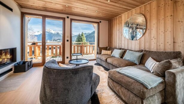 Ein gemütliches Wohnzimmer mit Kamin, einem großen Fenster mit Blick auf die Berge, einem runden Spiegel und einer Holzbalkendecke.