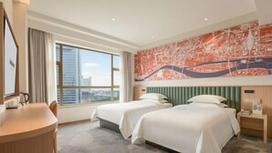 Quarto de hotel com duas camas, uma escrivaninha, uma cadeira, uma janela grande e vista para a cidade.
