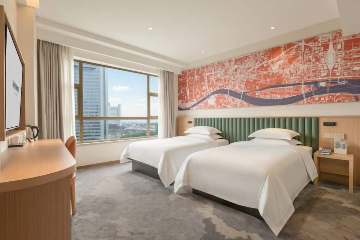Een hotelkamer met twee bedden, een bureau, een stoel en een groot raam, met uitzicht op de stad.