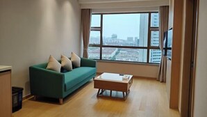 A modern living room with a green sofa, a wooden coffee table, and a large window offering a city view. - Guomao Boutique Hotel (Zhangjiagang)