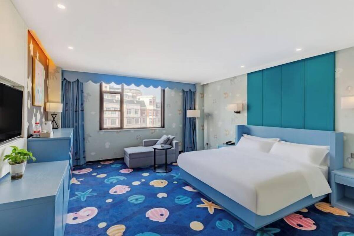 Quarto de hotel com cama, escrivaninha, cadeira, televisão, janela e vista para edifícios.