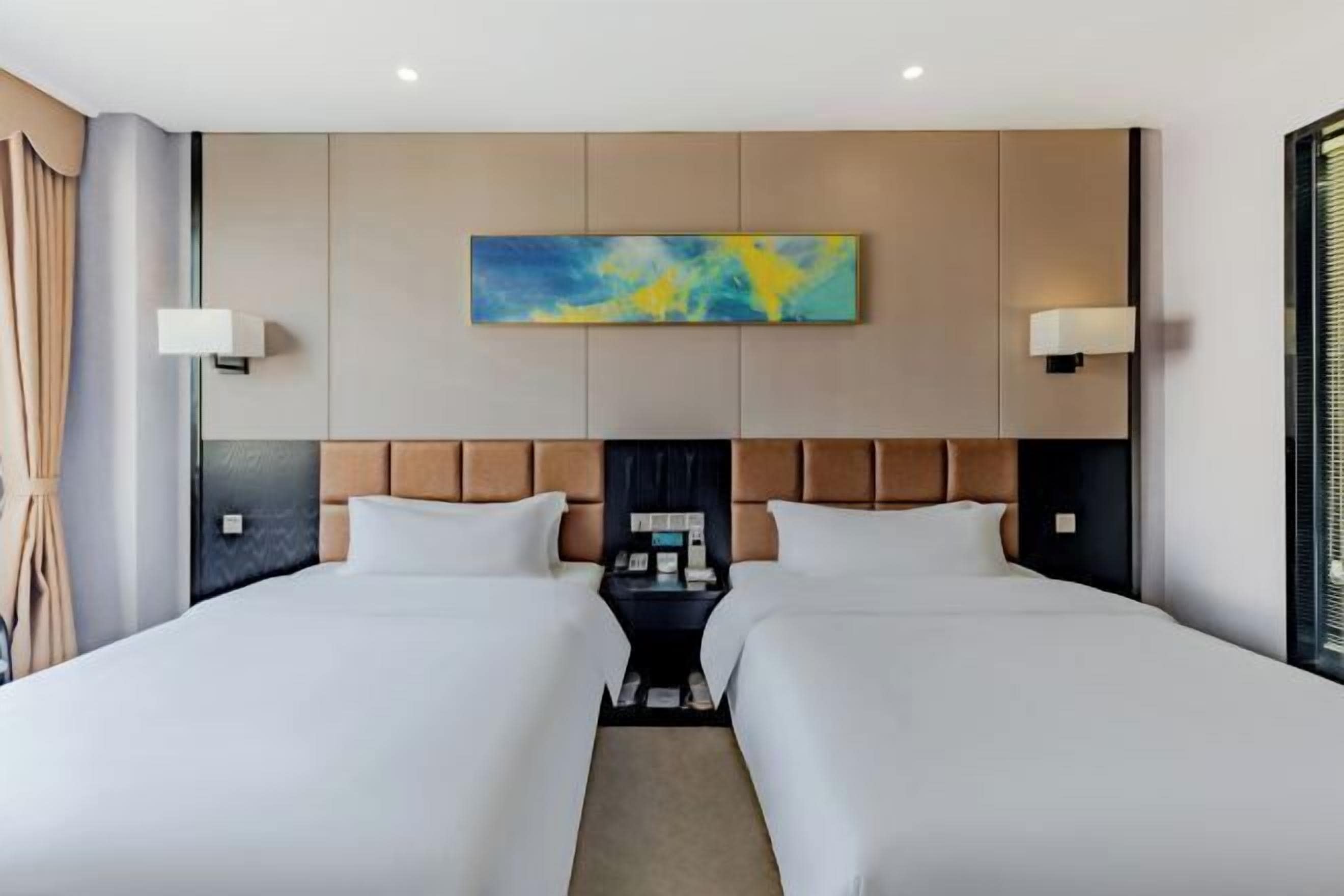 A hotel room with two beds, a headboard with a painting, bedside lamps, and a nightstand with a phone and books.