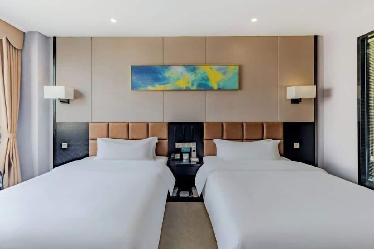 Quarto de hotel com duas camas, cabeceira com pintura, luminárias de cabeceira e mesa de apoio com telefone e livros.