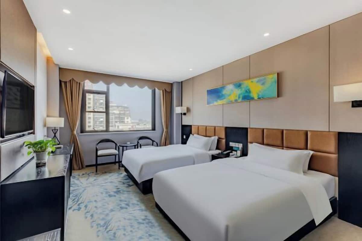 Quarto de hotel com duas camas, uma mesa de trabalho com televisão, uma planta e vista para a cidade.