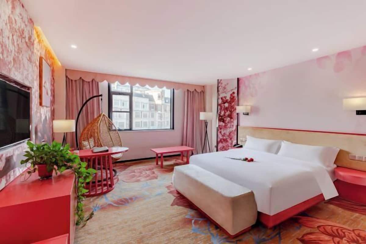 Quarto de hotel com uma cama, televisão, janela com cortinas, uma mesinha, uma cadeira e um vaso de plantas.