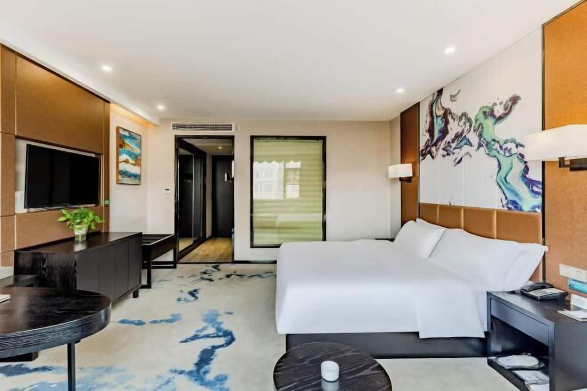A modern hotel room with a large bed, a desk, a TV, and a painting on the wall.
