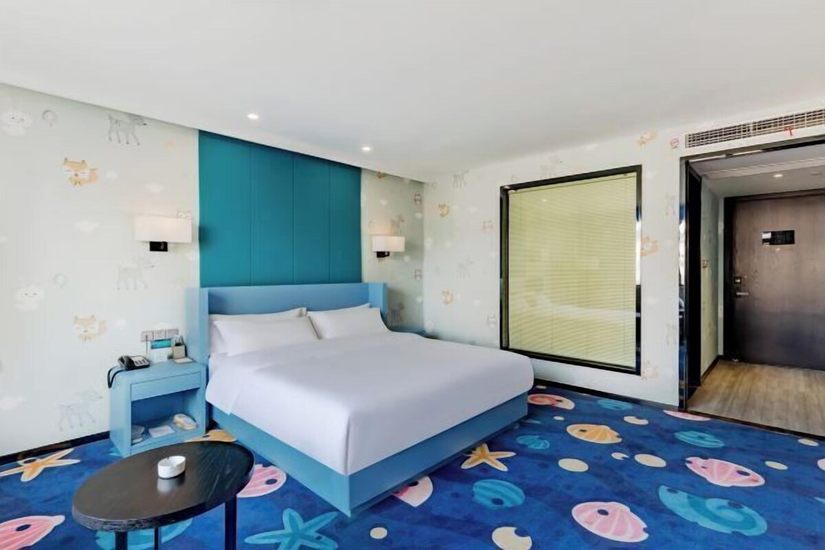 A hotel room with a bed, a nightstand, a small table, and a window with blinds.