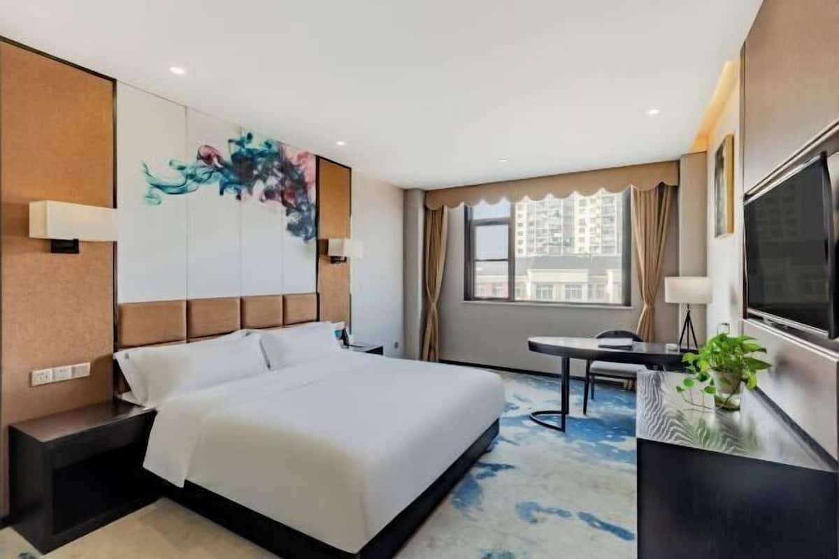 Quarto de hotel com cama, escrivaninha, televisão e janela com vista para a cidade.