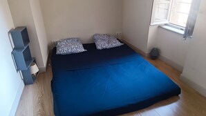 Une chambre à coucher avec un lit, deux coussins à motifs, une couvre-lit bleue, une étagère bleue, une lampe et une fenêtre.