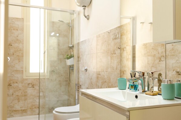 A modern bathroom with a glass shower enclosure, a white sink with teal accessories, and a marble-tiled wall. - Sardinia RE - Melany 22 (Alghero)
