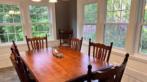 A dining room with a wooden table and chairs, large windows with a view of greenery, and a chandelier. - 3 Bedroom house, close to the beach and beech forest. (Eastham)