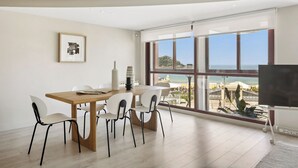 Un comedor moderno con una mesa de madera y sillas blancas, ventanas amplias con vista a la playa y un televisor de pantalla plana a la derecha.