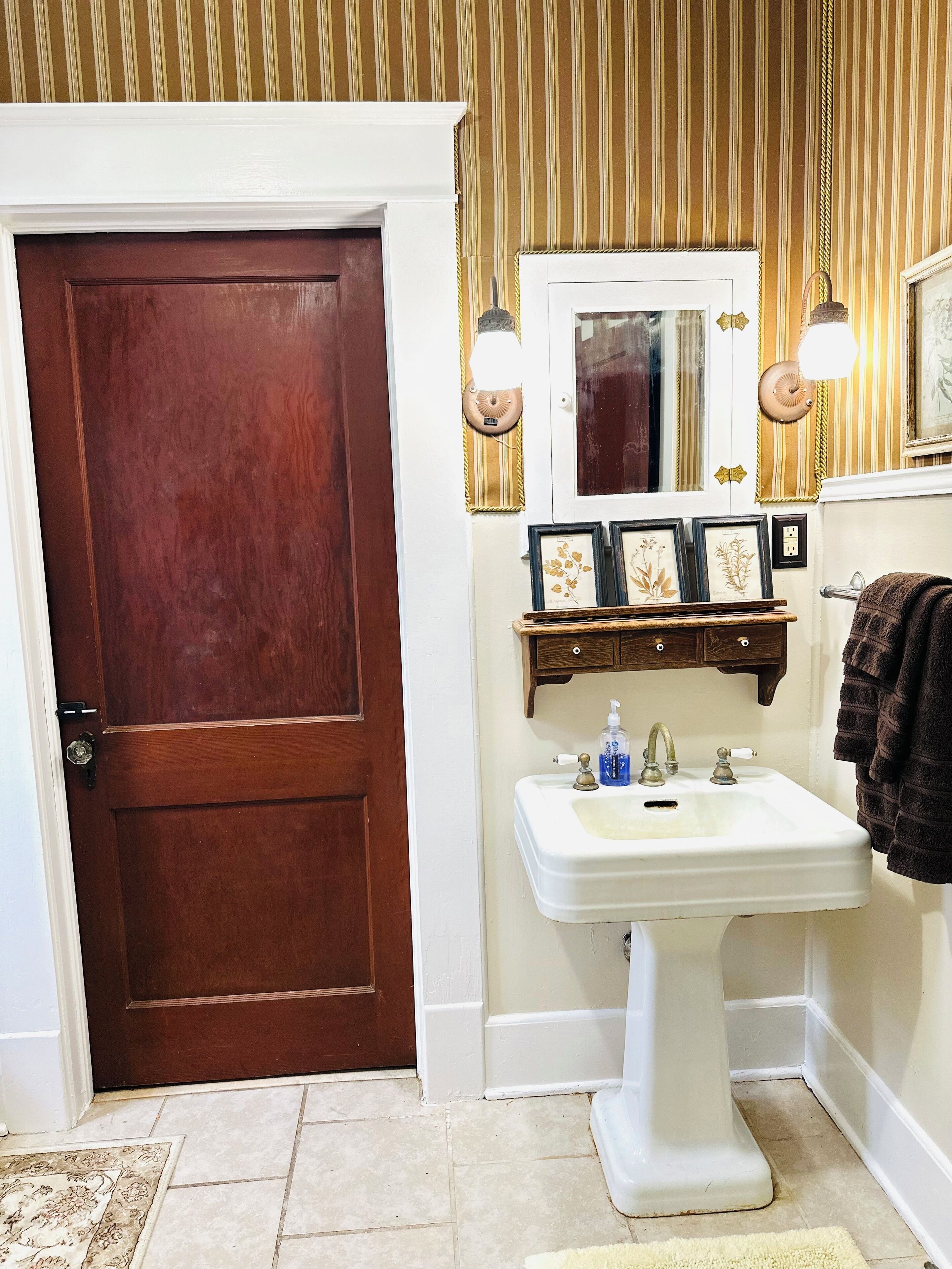 A bathroom with a wooden door, a white sink, and a mirror above it. There are framed pictures on the wall and a towel hanging on the right.