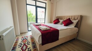 Un letto rifatto con cura, una coperta rossa e cuscini, un termosifone, un tappeto decorato e una finestra con vista sul verde.