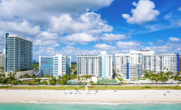 A beachfront hotel with multiple high-rise buildings, a sandy beach, and clear skies. - 2bd Oceanfront Master Ste OceanView w/balcony, Pool, nearby dining, beach access (Miami Beach)