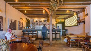 Een restaurantinterieur met houten tafels en stoelen, een bar met een bartender, en een bakstenen muur met ingelijste foto's.