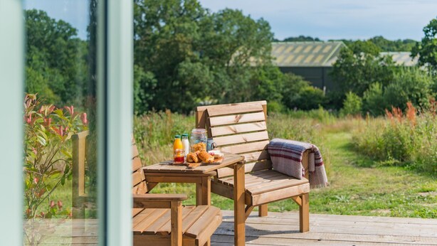 Een houten terras met een houten stoel en tafel, een plaid en ontbijtartikelen.