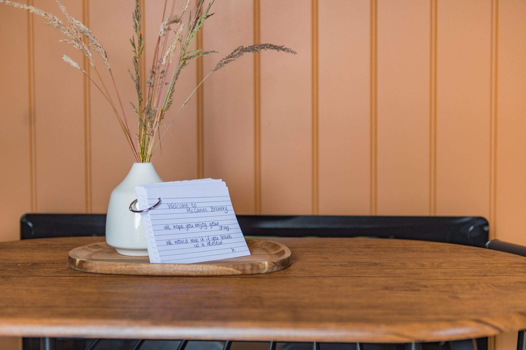 A wooden table with a white vase containing dried plants and a handwritten note.