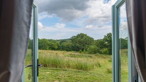Une porte ouverte donne sur une terrasse en bois, d’où l’on peut admirer un champ verdoyant et des arbres sous un ciel partiellement nuageux.