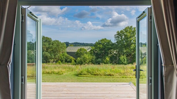 Een open deur die uitkomt op een houten terras met uitzicht op een groen landschap en bomen.