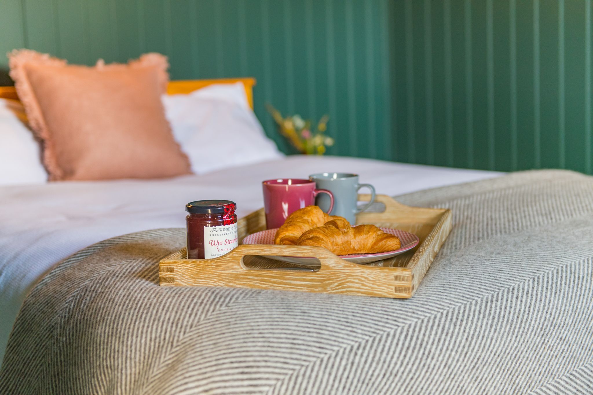 Bandeja de desayuno con croissants, un tarro de mermelada y dos tazas sobre una cama con cabecera verde y almohada rosa.