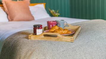Bandeja de desayuno con croissants, un tarro de mermelada y dos tazas sobre una cama con cabecera verde y almohada rosa.