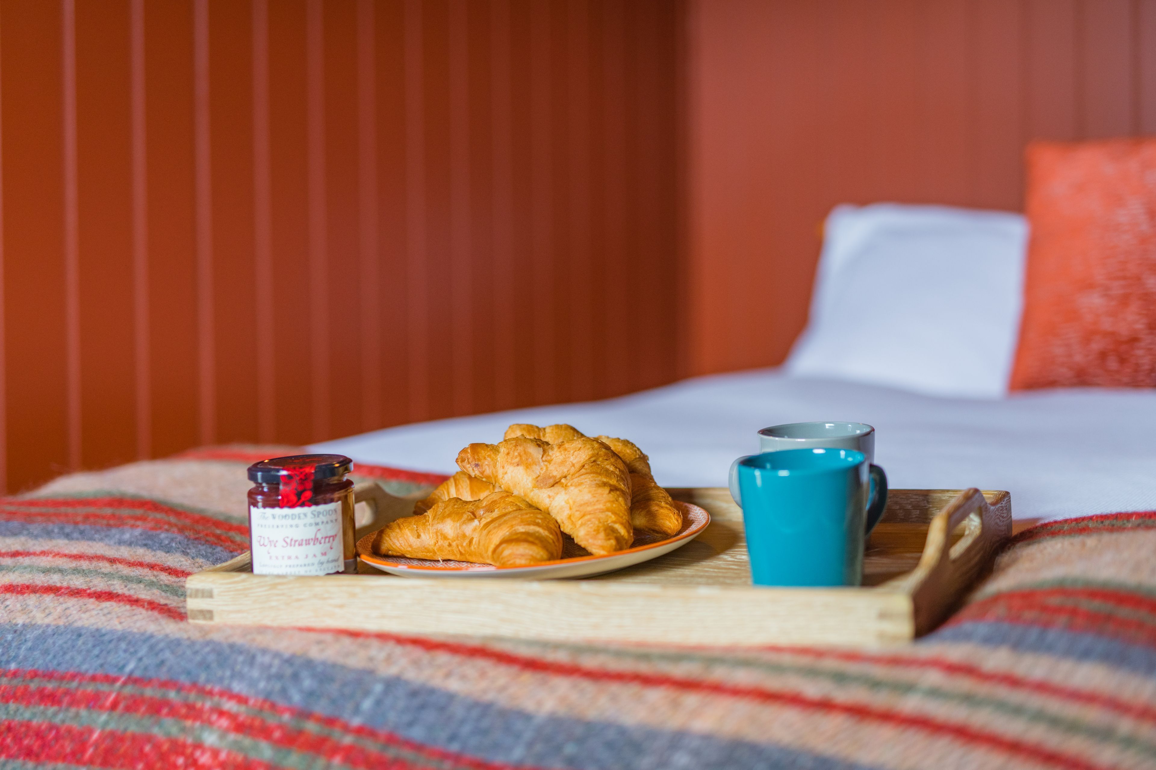 A bed with a striped blanket, a plate of croissants, a jar of jam, and a blue mug on a tray.