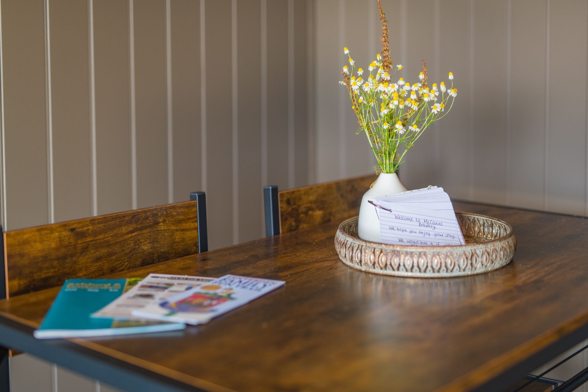 Ett träbord med en vit vas som innehåller gula blommor, en handskriven lapp och en tidning.