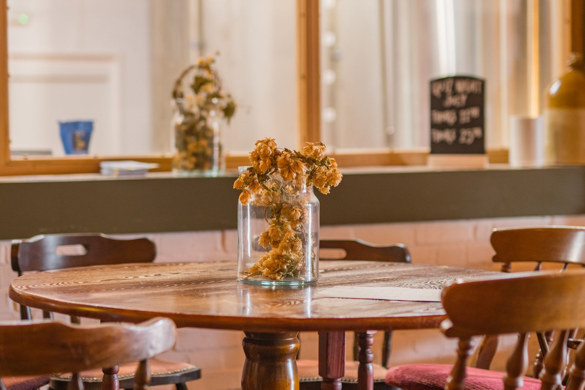 Een ronde houten tafel met een glazen vaas met gedroogde bloemen, omringd door houten stoelen.