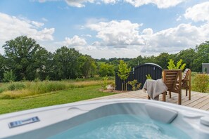 Un jacuzzi en una terraza de madera con vistas a un paisaje verde y un cobertizo negro al fondo.