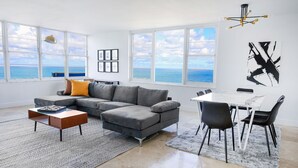 A modern living room with a gray sectional sofa, a coffee table, a dining table with chairs, and a view of the sea through large windows. - 2bd Oceanfront Master Ste OceanView w/balcony, Pool, nearby dining, beach access (Miami Beach)