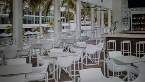 A bar area with white bar stools and tables, large windows with views of palm trees, and a stone wall with a mounted TV. - Charming 3-bedroom Gulf-view villa with many amenities in amazing Marco Island (Marco Island)