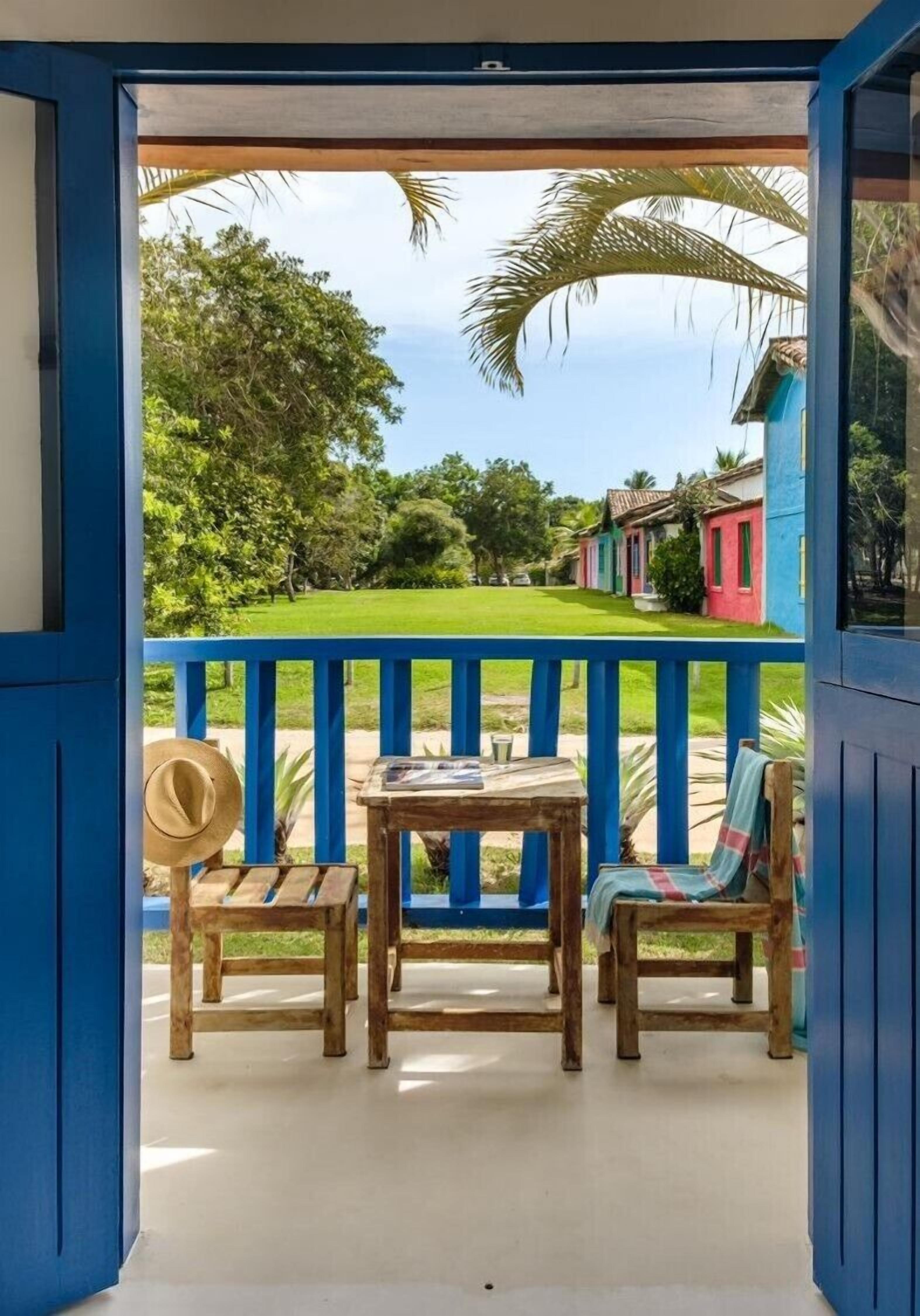 Varanda com mesa e cadeiras de madeira, um chapéu e vista para casas coloridas e vegetação.