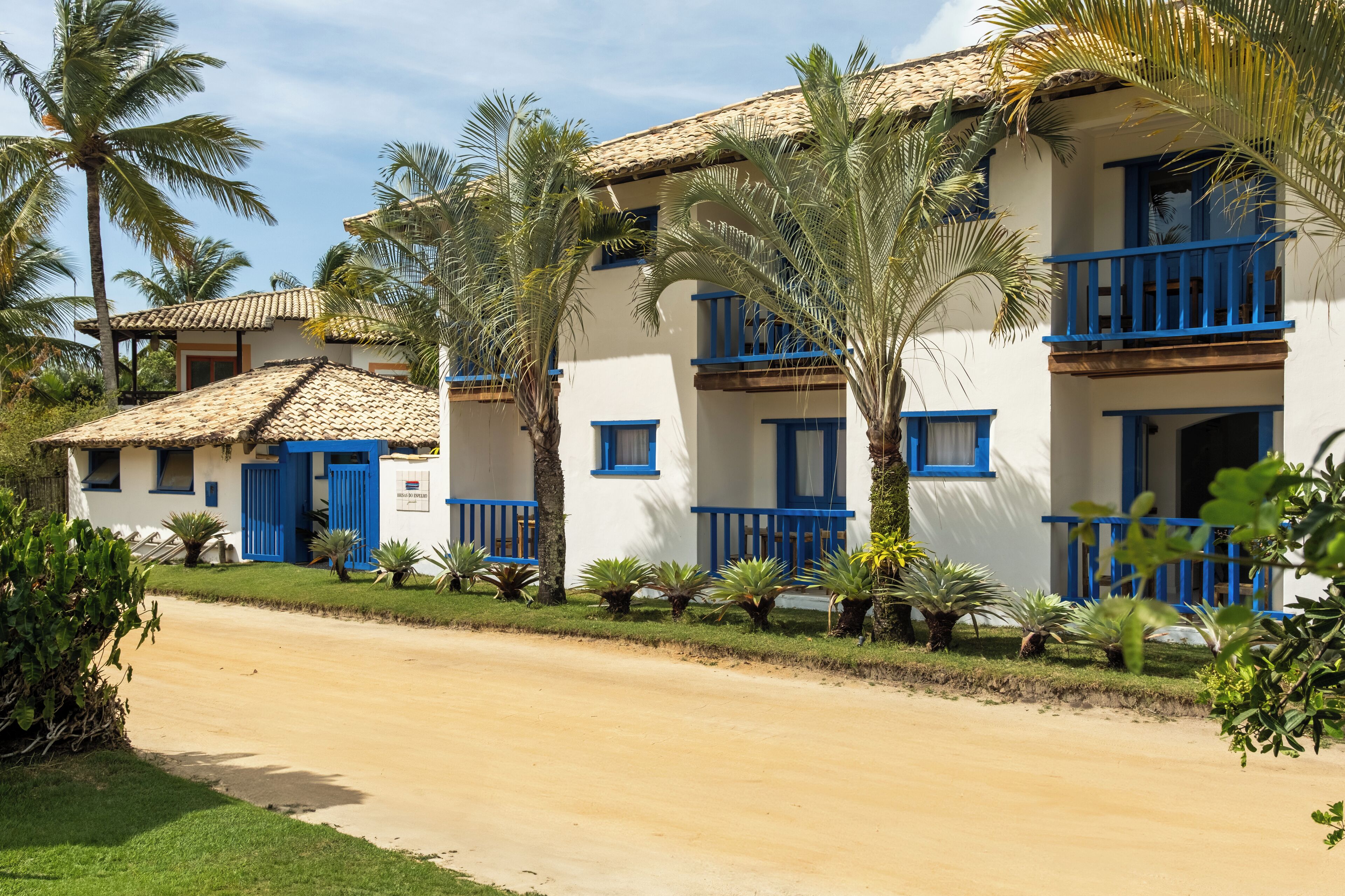 Una hilera de edificios blancos con balcones y puertas azules, rodeada de palmeras y plantas.