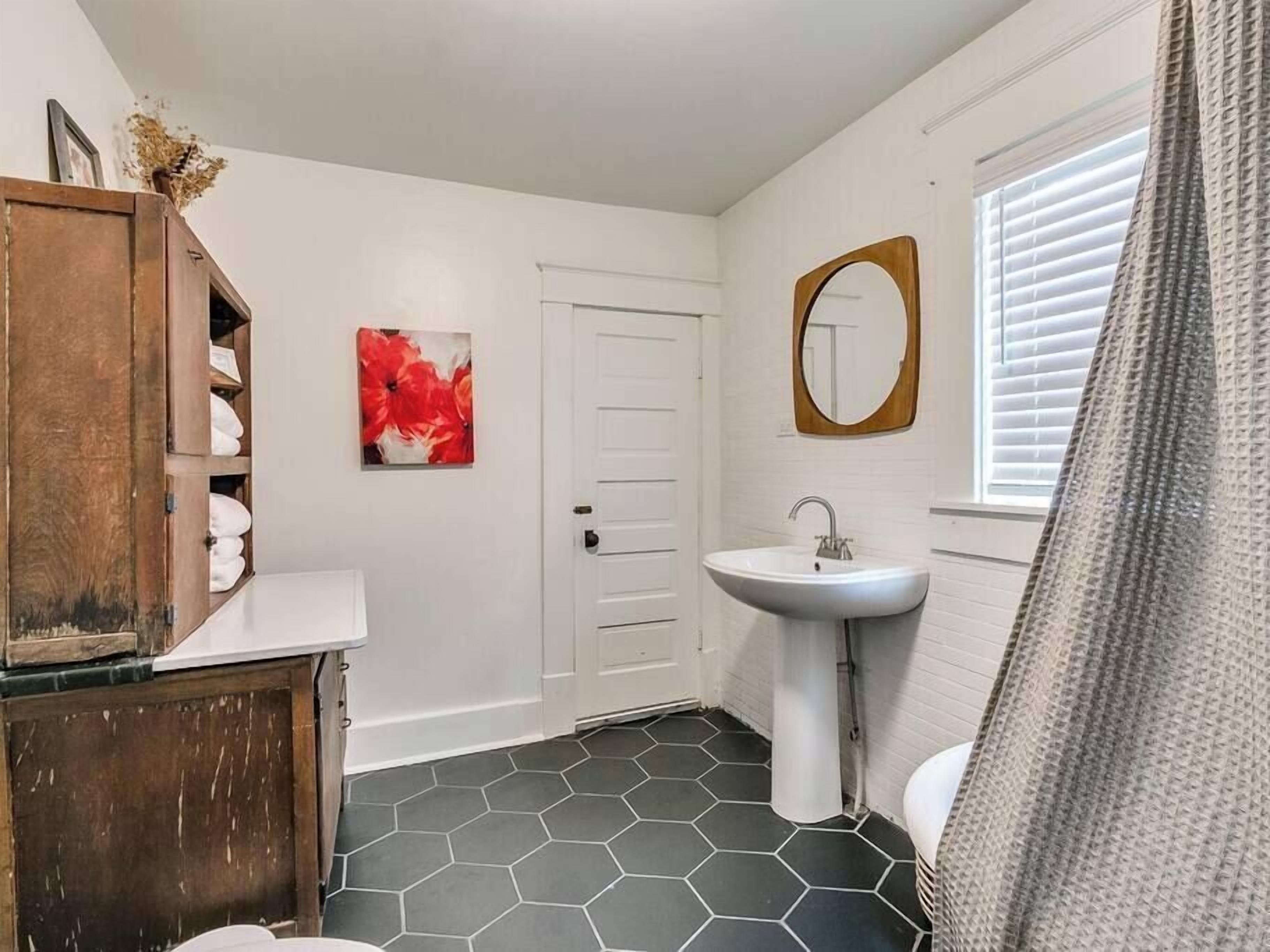 A bathroom with a hexagonal tile floor, a white sink, a wooden cabinet with towels, a red floral artwork, and a white door.