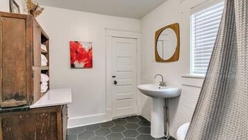 A bathroom with a hexagonal tile floor, a white sink, a wooden cabinet with towels, a red floral artwork, and a white door.