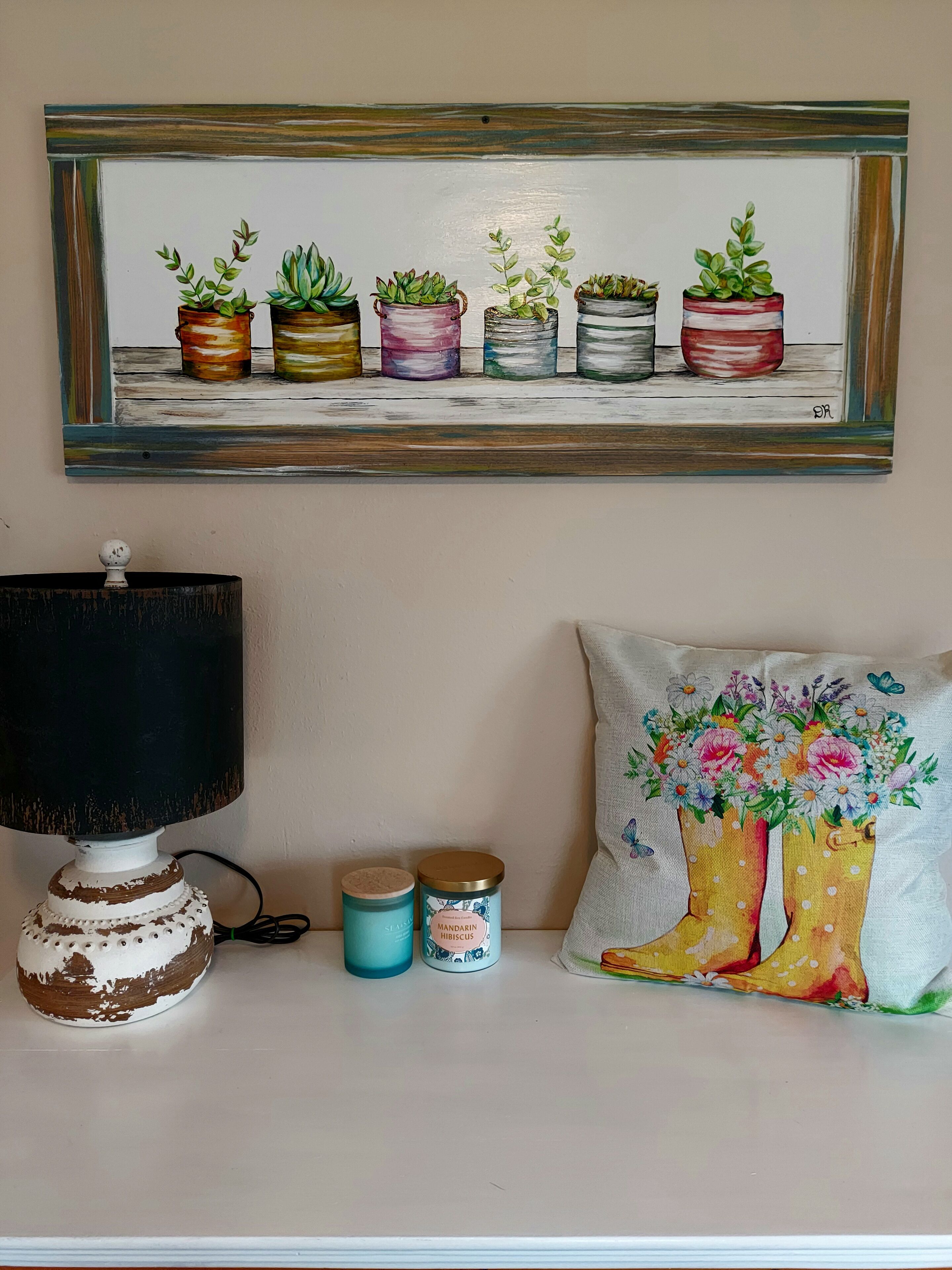 A wall with a framed painting of potted plants, a lamp, and decorative items including a pillow and two containers.