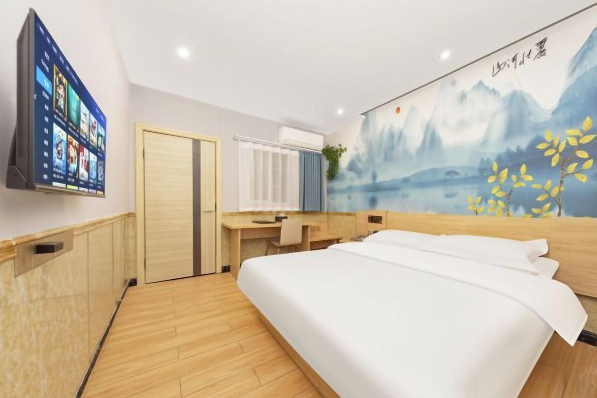 A hotel room with a bed, a desk, a TV, and a wall with a mountain painting.