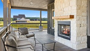 Un área de estar al aire libre techada con chimenea de piedra, terraza de madera y vista a una casa a lo lejos.