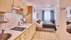 Habitación de hotel pequeña con kitchenette, cama con colcha oscura y vista de la ciudad por la ventana.