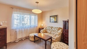 Una sala de estar con un sofá, una mesa de centro de madera, una mesita de noche con una lámpara, una ventana con cortinas traslúcidas y un cuadro enmarcado en la pared.