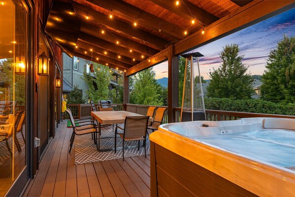 Una terraza de madera con un jacuzzi, asientos al aire libre y luces colgantes.