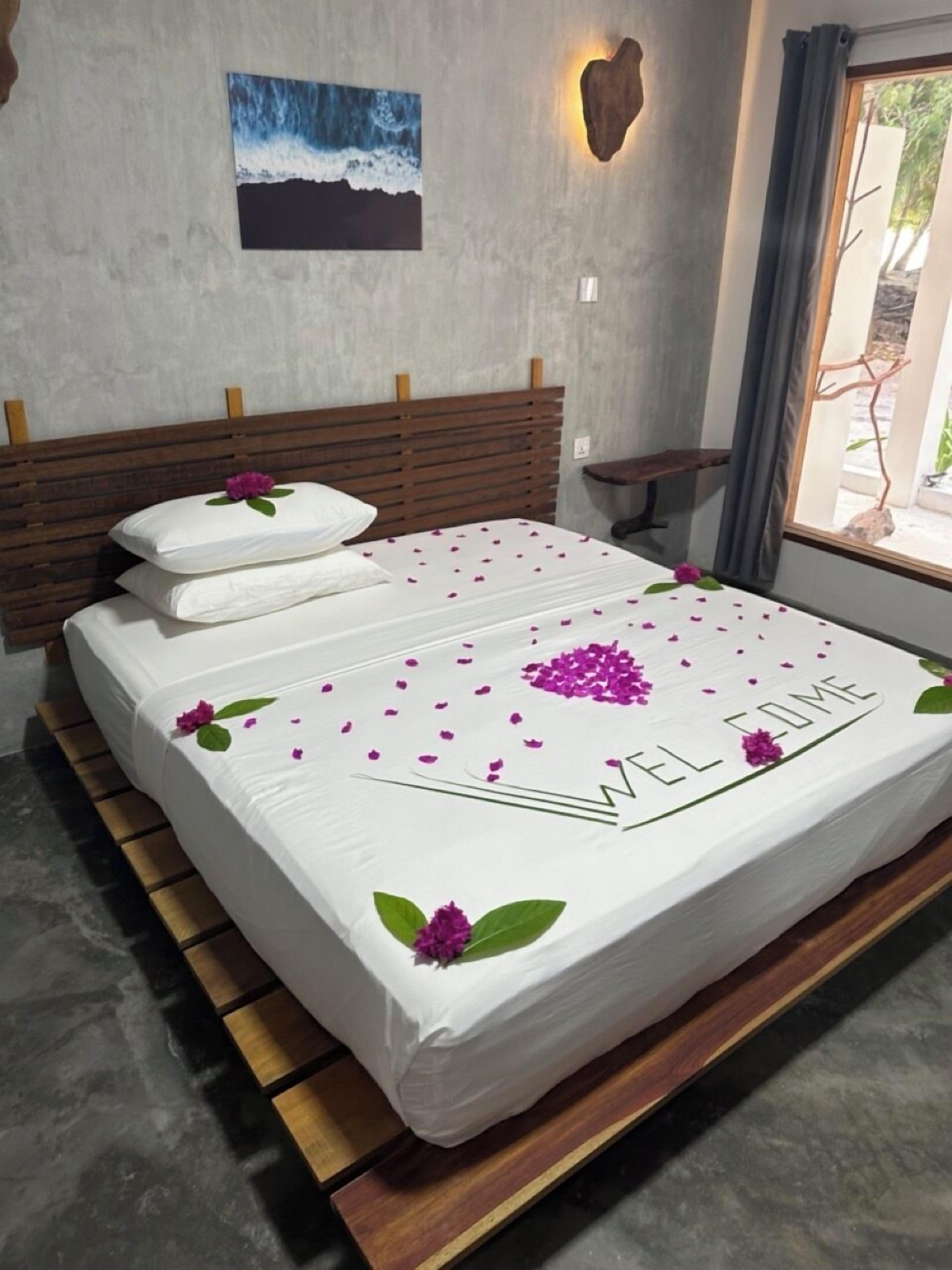 A neatly made bed with a 'WELCOME' sign, wooden headboard, and a small bench beside the window.