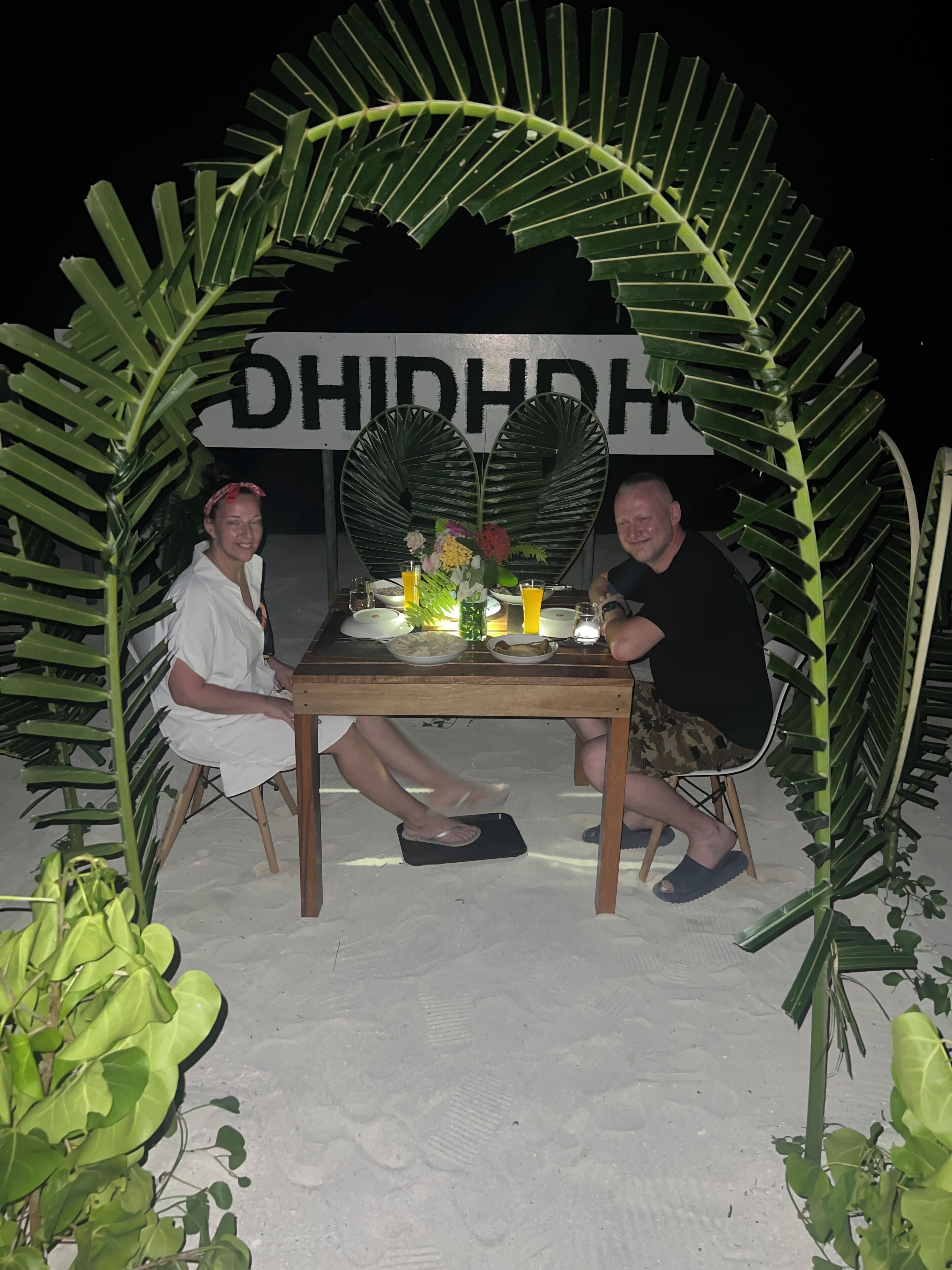 Dos personas sentadas en una mesa con un paisaje tropical, con un cartel que muestra el texto "DHIDHRH".