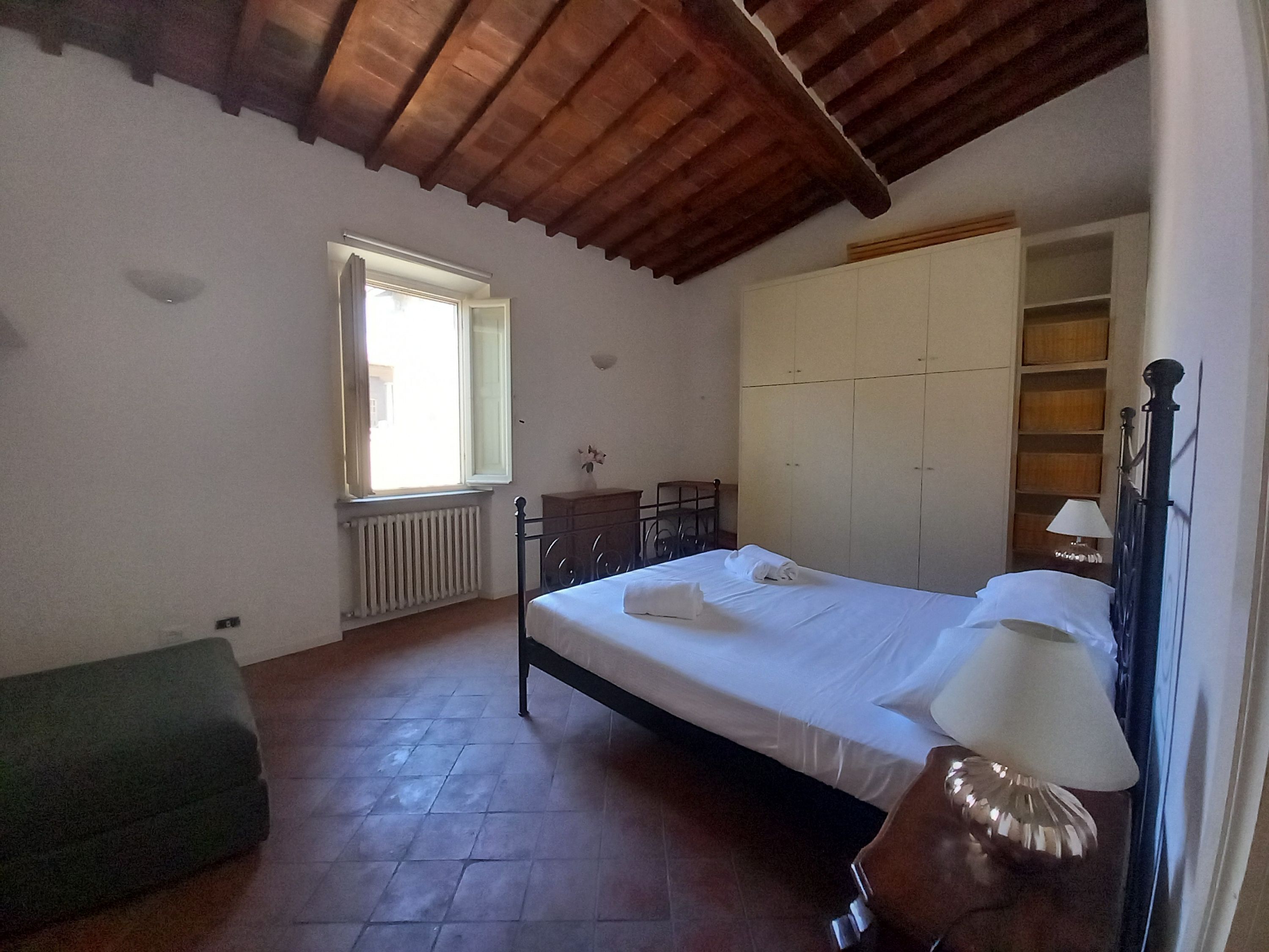 A bedroom with a bed, a radiator, a window, a wooden wardrobe, and a green bench.