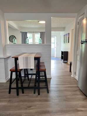 A kitchen with a wooden table and two stools, a refrigerator, and a window with curtains.
