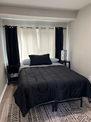 A bedroom with a bed, black curtains, a bedside table, and a patterned rug.