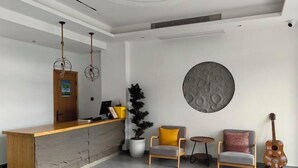 Ένας χώρος υποδοχής με ένα ξύλινο γραφείο, δύο καρέκλες, ένα μικρό τραπέζι, ένα φυτό σε γλάστρα και μια κιθάρα.