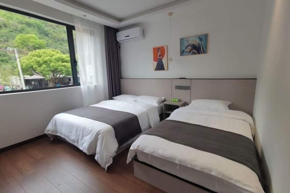 A hotel room with two beds, a large window with a view of greenery, an air conditioning unit, and a painting on the wall.