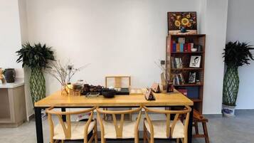 A dining area with a wooden table and chairs, a bookshelf with books and a painting, and two potted plants.